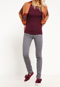 Vínový top s krajkovými detaily, šedé přiléhavé kalhoty a bordó tenisky s mašlemi. Modelka má na sobě oranžovo-šedý proužkovaný kardigan.