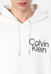 Balta medvilnė trumpomis rankovėmis su raišteliais, priekyje esantis juodas "Calvin Klein" logotipas. Minkšta tekstūra ir kasdieniškas dizainas.