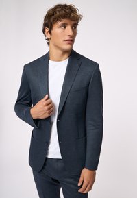 Dunkelblazer aus strukturiertem Stoff, einreihiges Design mit Reverskragen, zwei vordere Taschen und darüber ein schlichtes weißes T-Shirt.