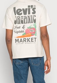 Krémové bavlněné tričko s barevným potiskem na přední straně, zobrazující zeleninu a velkým textem "Levi's Organic Fruit & Vegetable Market."