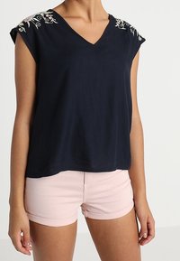Mörkblå V-ringad topp med blommig broderi på axlarna, gjord i lättviktsmaterial, som matchas med rosa shorts med rullad kant.