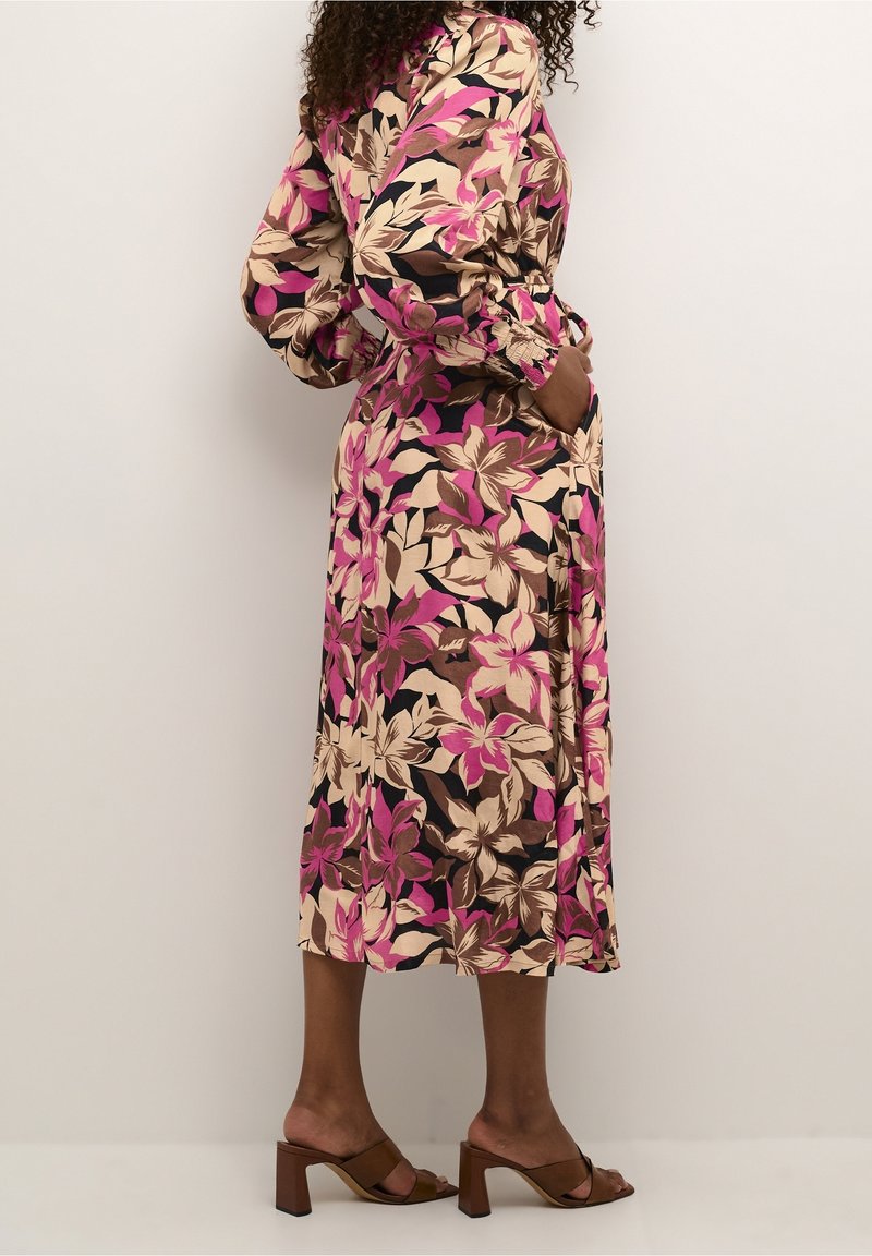 Femme portant une robe longue à manches longues, longueur cheville, à fleurs dans des tons rose, beige et marron, avec des sandales à talons marron, debout de profil, les mains dans les poches.