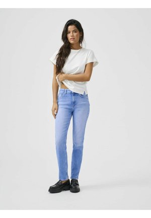 Kobieta z długimi ciemnymi włosami, ubrana w biały krótki rękaw, jasnoniebieskie dżinsy i czarne buty, stojąca na tle jednolitego szarego tła.