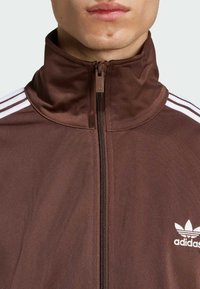 Veste zippée marron avec col haut, comportant des rayures blanches sur les épaules et un petit logo Adidas blanc sur le côté gauche. Texture lisse.