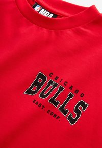 Crvena pamučna majica s crnim printom "CHICAGO BULLS" i "EAST. CONF." u podebljanom, koledž-stilu fonta. Okrugli izrez, kratki rukavi.