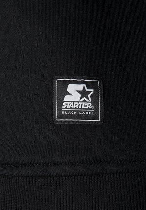 Material negru cu textură ondulată; prezintă un eticheta dreptunghiulară alb-negru cu textul "STARTER BLACK LABEL" tipărit cu litere îngroșate.
