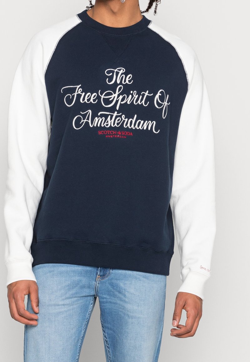 Marineblaues Sweatshirt mit weißen Raglanärmeln, ausgestattet mit aufgesticktem Text "Der Freigeist von Amsterdam" und einem roten Logo.