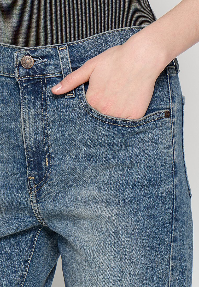 Los jeans de mezclilla azul presentan un diseño clásico con un bolsillo frontal, costuras visibles y un botón de metal en la cintura. La textura del textil es suave.