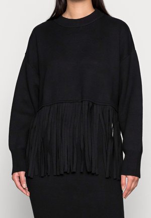 Pull noir à manches longues avec ourlet à franges porté par une personne debout devant un fond clair uni.