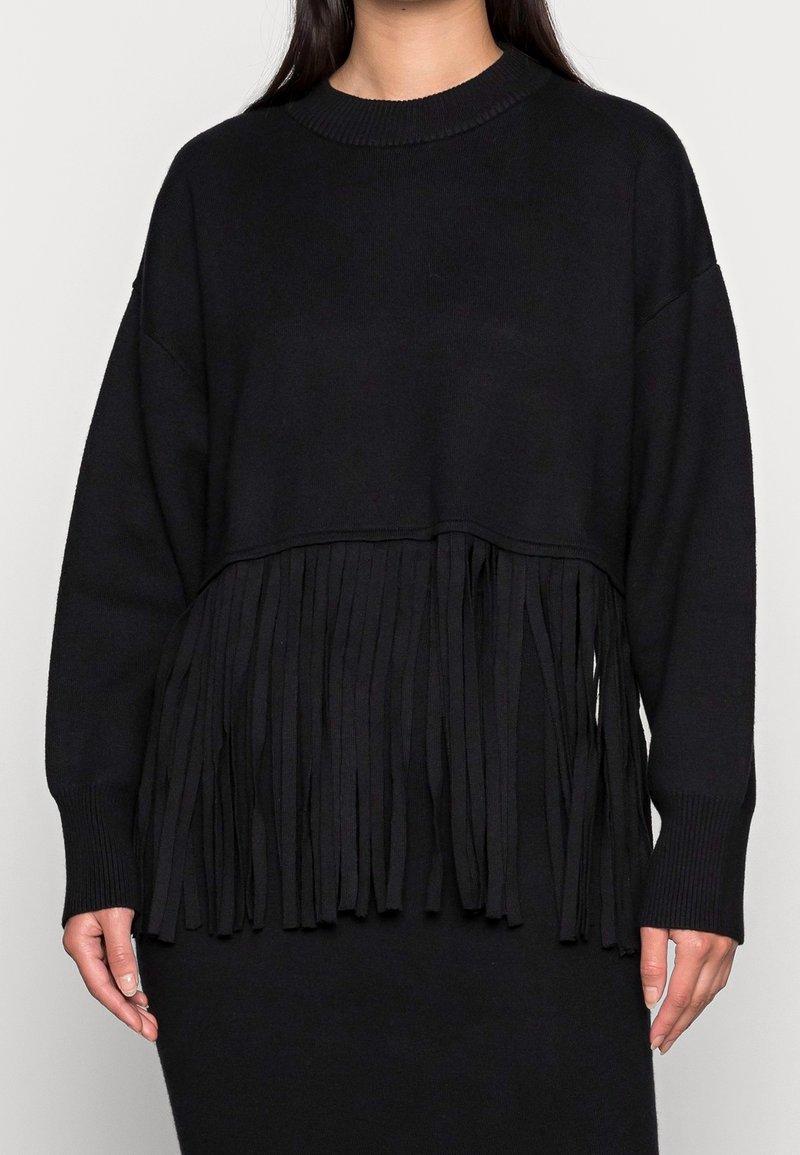 Pull noir à manches longues avec ourlet à franges porté par une personne debout devant un fond clair uni.