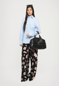 Geantă de mână din piele neagră cu un design texturat, purtată cu o cămașă bleu deschis și pantaloni largi cu un model colorat reprezentând ceaiuri și deserturi.