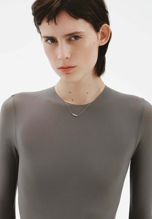 Top gris de manga larga hecho de un material suave y elástico. El escote es redondo. Se lleva un delicado collar dorado con un colgante curvado.