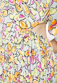 Robe fleurie avec des fleurs orange, jaune et rose, avec une taille froncée, une découpe latérale et un détail de nœud en tissu noué.