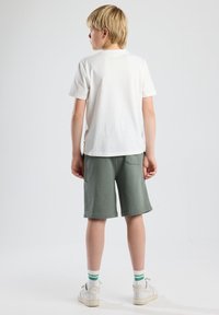 T-shirt bianco in cotone abbinato a pantaloni corti verde oliva. I pantaloni corti presentano una tasca posteriore. Modello che indossa sneakers bianche con calzini a righe verdi.