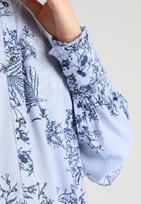 Vaaleansininen nappikauluspaita, jossa on tummansinisestä kukkakuviosta, teksturoitu pinta ja kerätty hihansuu.