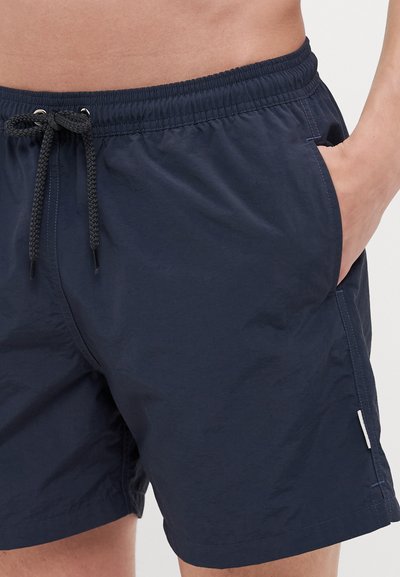 Marineblå svømmebukser laget av lett stoff, med strikk i livet og snor, sidelommer og en liten logoetikett.