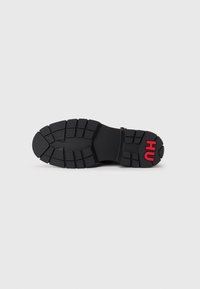 Suela de zapatilla de goma negra con un patrón de relieve prominente. Presenta el logo rojo "HU" en el talón exterior. Diseñada para ofrecer tracción y durabilidad.