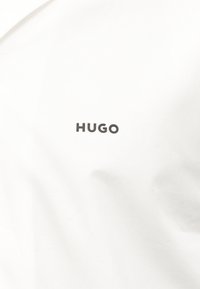 Λευκό ύφασμα με μαύρο κείμενο στο κέντρο που γράφει "HUGO" με έντονα κεφαλαία γράμματα.