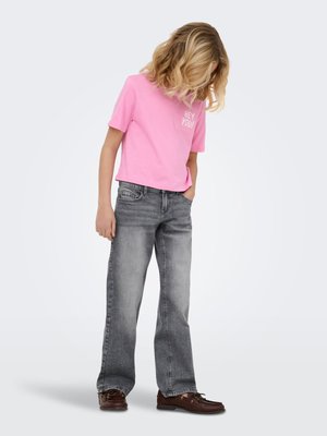Enfant aux cheveux blonds bouclés portant un t-shirt rose "HEY YOU!", un jean gris et des chaussures marron, regardant vers le bas avec une main dans la poche.