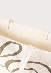 Τσάντα από βαμβάκι σε κρέμα χρώμα με σκούρο πράσινο ακαθόριστο σχέδιο. Διαθέτει ανθεκτικούς χειρολαβές και ένα απλό, ακατάλληλο εσωτερικό.