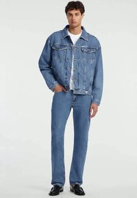 Ung man som bär en blå denimjacka och matchande jeans över en vit skjorta, står med ena handen i fickan och svarta skor.