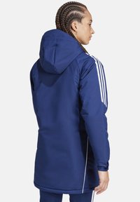 Marineblauer, wasserdichter Parka mit Kapuze, ausgestattet mit weißen Streifen an den Ärmeln und einem geraden Schnitt. Glatte Stoffstruktur, bis zur Mitte des Oberschenkels.