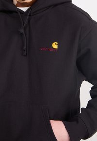 Felpa nera con cappuccio e tasca frontale. I dettagli distintivi includono un logo ricamato giallo e marrone. Tessuto morbido.