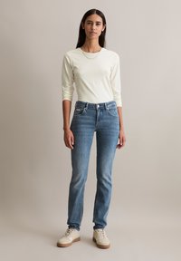 Jasna kremowa bluzka z długim rękawem, niebieskie dżinsy o wyblakłym kolorze i beżowe trampki. Prosty design, standardowy krój i brak widocznych wzorów.