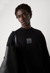 Svart hettegenser med rund hals og grå firkantet logo. Modellen holder en svart lær totebag med en glatt tekstur og buet form.