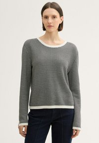 Schwarzer und weißer gestrickter Pullover mit strukturiertem Muster, gerippten Bündchen und rundem Halsausschnitt, kombiniert mit dunkelblauen Hosen.