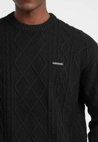 Maglione nero a maglia intrecciata con scollo rotondo e motivi a diamante testurizzati. Presenta un piccolo logo "GUESS JEANS" sul davanti.