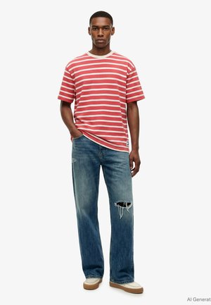 Männliches Model trägt ein rot-weiß gestreiftes T-Shirt, blaue zerrissene Jeans und beige Sneakers, steht mit einer Hand in der Tasche.