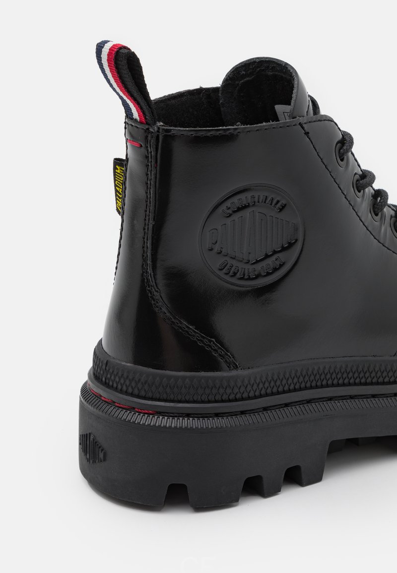Bottine en cuir verni noir avec une semelle en caoutchouc texturée, arborant un logo circulaire, des accents rouges, et une languette rayée.