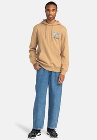 Tan hooded sweatshirt with front pocket and blue graphic detailing. Wide-leg blue denim jeans and black athletic shoes complete the look.