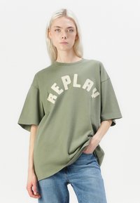Olijfgroene oversized t-shirt met korte mouwen, voorzien van "REPLAY" in vet crèmekleurige letters op de borst. Zacht katoenmateriaal.