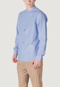 Camicia azzurra chiara con bottoni, realizzata in tessuto testurizzato, con colletto a fascia e bottoni colorati. Abbinata a pantaloni a strisce marroni.