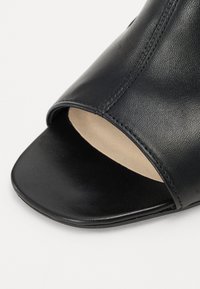 Chaussure noire en cuir à bout ouvert avec une texture lisse, une forme arrondie et des accents cousus sur le bord supérieur.