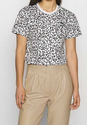 Kvinde iført hvid leopardprint kortærmet skjorte indstukket i beige højtaljede plisserede bukser, stående mod en neutral baggrund.