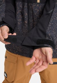 Handen die de zwarte klittenbandmanchet van een gevlekte zwart-bruine jas aanpassen, met daaronder bruine broek zichtbaar tegen een witte achtergrond.