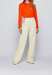 Röd långärmad blus med plisserade detaljer, tillsammans med högmidjade, vida vita byxor med guldklickar och slätt tyg.