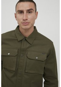 Camisa chaqueta de algodón verde oliva con dos bolsillos en el pecho, botones a presión y un cuello clásico. Textura suave con costuras en contraste.