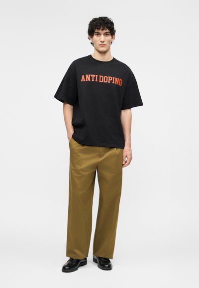Jeune homme debout les mains dans les poches, portant un t-shirt noir "ANTI DOPING", un pantalon marron à jambes larges et des chaussures noires sur fond uni.