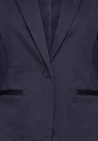 Blazer bleu marine en tissu texturé, doté d'une fermeture à un bouton, de revers crantés et de deux poches avant avec des accents noirs.