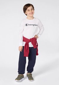 Mosolygó fiú, fehér hosszú ujjú Champion pólót, tengerészkék melegítőt, olajzöld sportcipőt viselve, piros pulóvert megkötve a derekán, fehér háttér előtt áll.