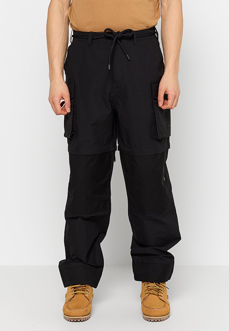 Pantalons cargo noirs avec taille ajustable, deux poches cargo sur les côtés et zone des genoux renforcée, associés à des bottes de travail beige.