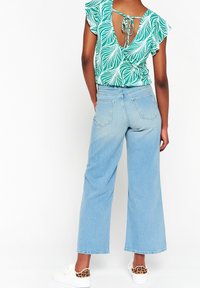 Zielono-biała bluzka w wzory z falbaniastymi rękawami i wiązaniem z tyłu, zestawiona z jasnoniebieskimi spodniami typu wide-leg i białymi sneakersami z lwimi akcentami.