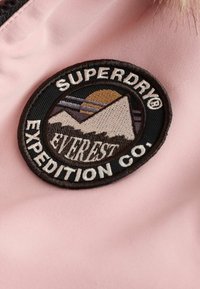 Parche de logotipo bordado en tela rosa, con un diseño de montaña, sol y el texto "SUPERDRY® EVEREST EXPEDITION CO." en marrón oscuro y blanco.