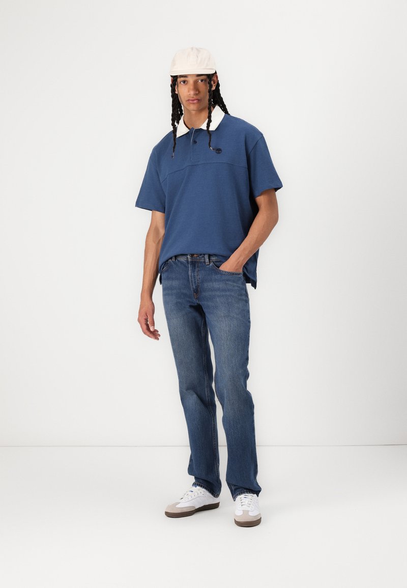 Polo shirt bleu à manches courtes avec col blanc, associé à un jean bleu et des baskets blanches. Le modèle se tient debout avec une main dans la poche.
