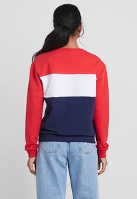 Femme aux cheveux foncés attachés en queue de cheval, portant un sweat-shirt bicolore rouge, blanc et bleu marine, et un jean bleu clair, tournée de dos vers la caméra.