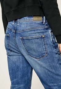 Blue denim jeans with a fitted design, featuring stitched back pockets, a leather patch on the waistband, and a slight fade in the fabric.